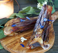 Load image into Gallery viewer, Two lamb shanks on a wooden board, bone-in and roasted to golden brown with blue flower petals