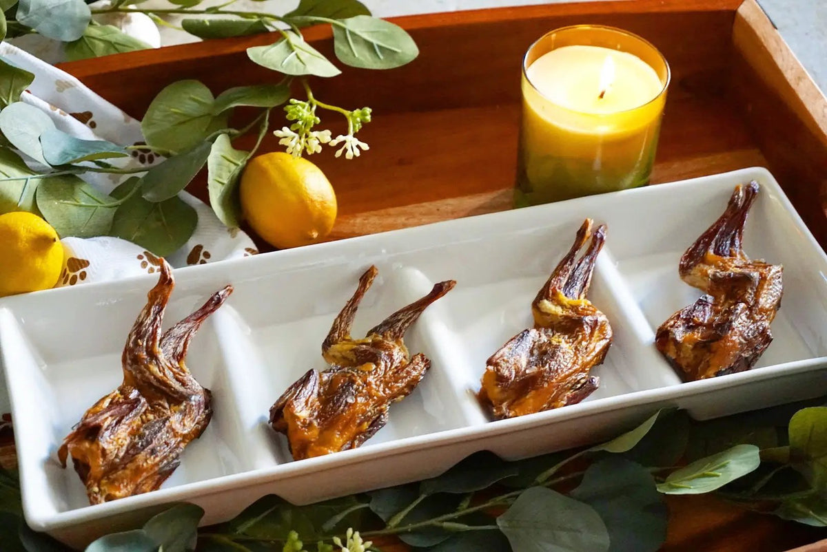 Whole quails (bone-in) by Naked Beasts, four roasted birds on a white serving tray with lemons and greenery.