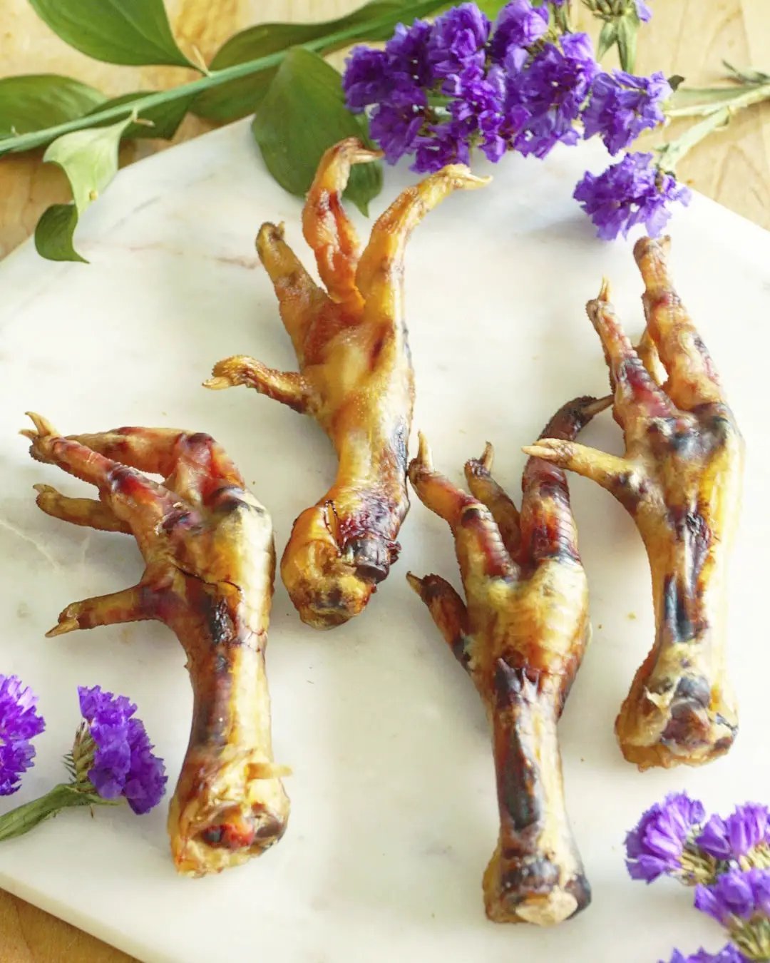 Chicken feet, four golden-brown roasted on a white marble board with purple flowers.