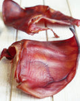 Carica l'immagine nel visualizzatore della galleria, Pig Ears by Naked Beasts; two dried reddish pig ears on a light wooden surface.
