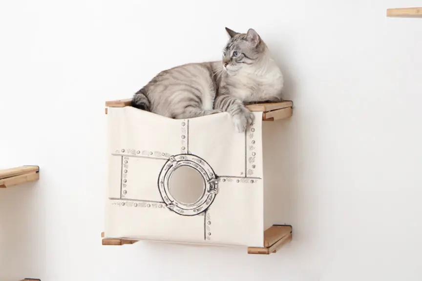 Enclosed Cat Bed - Cat Wall Cubby in beige canvas with riveted circular porthole; cat resting on top.