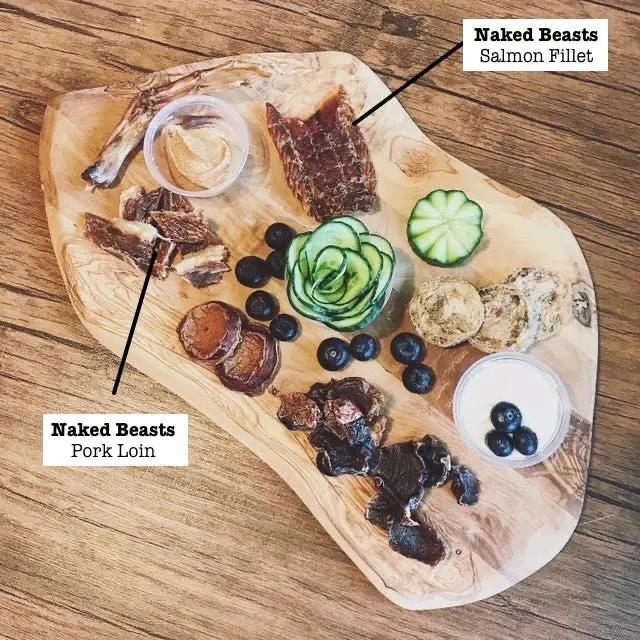 Salmon Fillet and Pork Loin from Naked Beasts on a wooden board with blueberries and cucumber garnish.