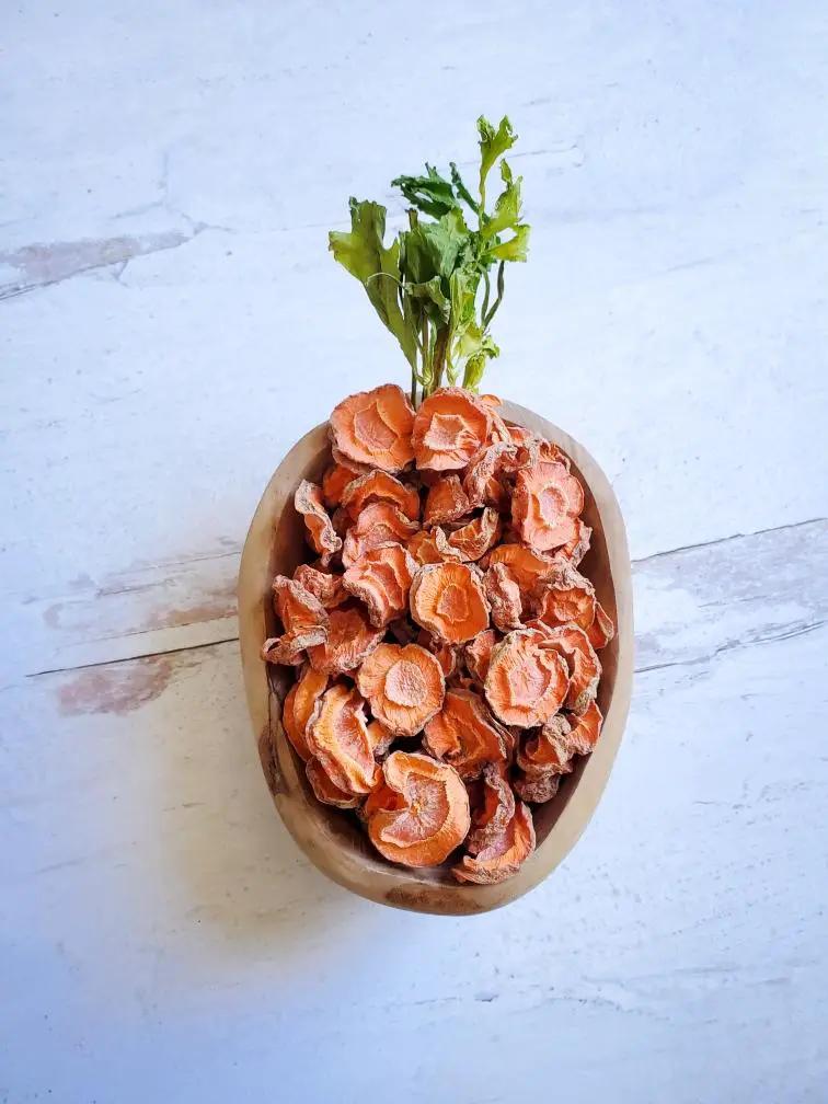 Carrots by Naked Beasts: orange dried carrot slices in a wooden bowl with green stems.