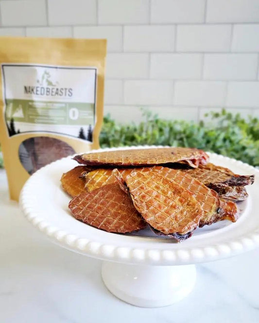 Bull Fries by Naked Beasts five golden-brown beef jerky slices on a white pedestal plate with brand bag visible behind.