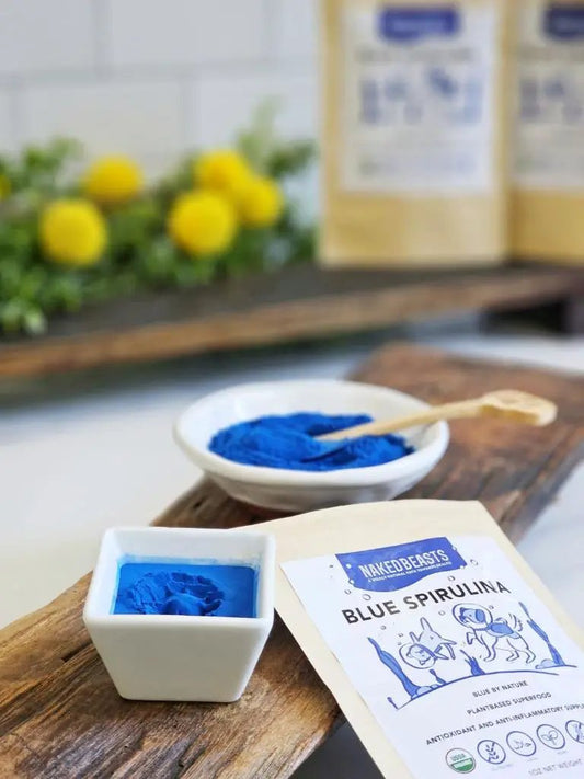 Blue spirulina powder (organic) by Naked Beasts in two white bowls on wood, vivid blue powder with a wooden spoon.