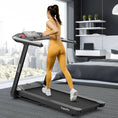 Carica l'immagine nel visualizzatore della galleria, Black treadmill with red buttons; woman in mustard activewear running in a modern room with city view; SuperFit

