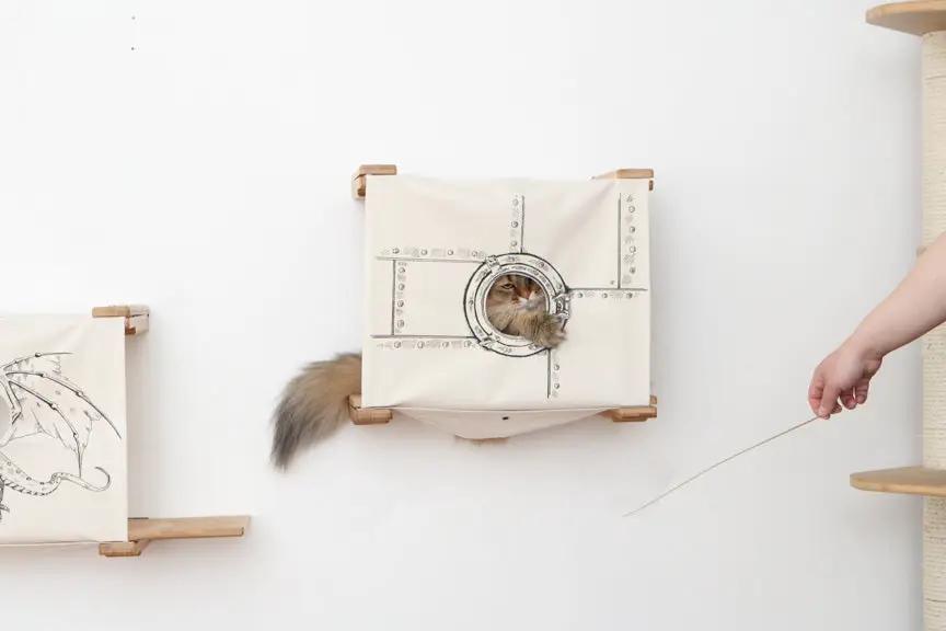 Enclosed cat bed wall cubby in beige fabric with circular doorway and riveted metal panel; two cubbies